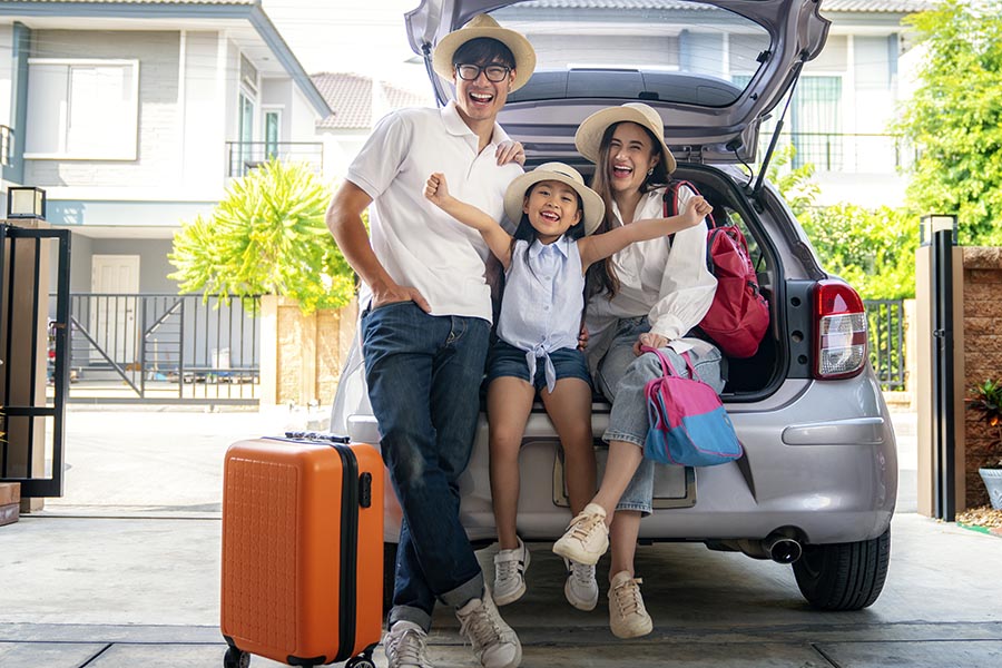 About Our Agency - Mother, Father and Young Daughter Sit In the Back Hatch of Their Car, Packed for a Vacation